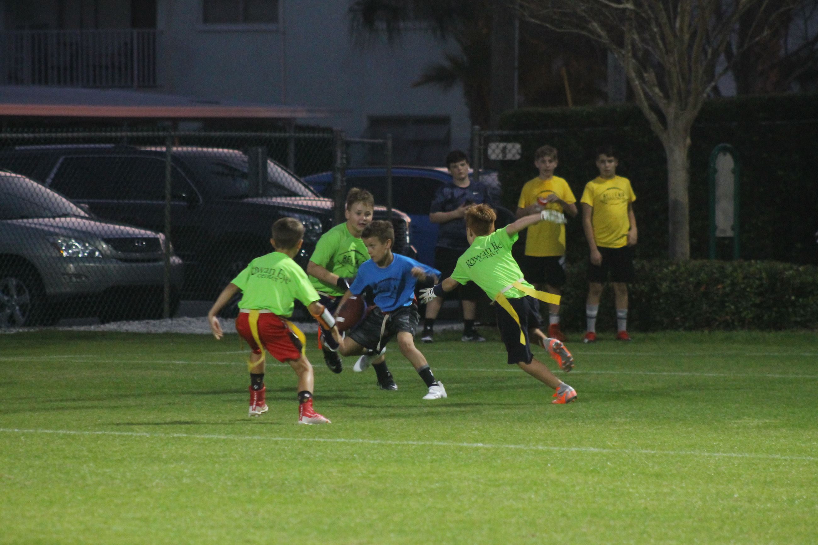 One boy holds a football while three surround him, attempting to pull his flag football flag 